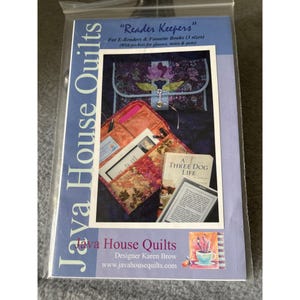 May include: A package for "Reader Keepers" by Java House Quilts. The package shows a quilted case with pockets for e-readers and books. The case is in shades of purple, orange, and floral patterns. The text on the package reads "For E-Readers & Favorite Books (3 sizes)".