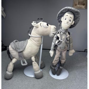 K&ouml;nnte beinhalten: Ein Pl&uuml;schtiersatz mit einer Cowboy-Puppe und einem Pferd. Die Cowboy-Puppe tr&auml;gt einen Hut, eine Weste und Stiefel. Das Pferd hat einen Sattel und wird von einem St&auml;nder gehalten. Beide Spielzeuge sind haupts&auml;chlich grau und beige.