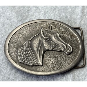 May include: Oval-shaped silver-toned metal belt buckle featuring a detailed relief of a horse's head. The horse's mane and facial features are intricately rendered. The buckle has a textured background and a rectangular bar for the belt.