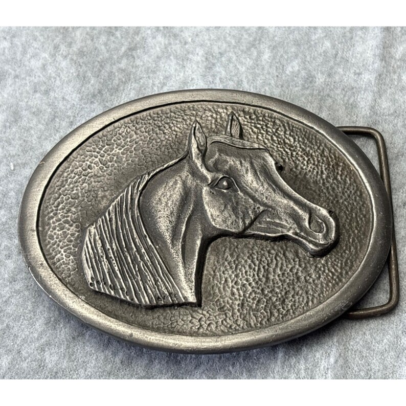 May include: Oval-shaped silver-toned metal belt buckle featuring a detailed relief of a horse's head. The horse's mane and facial features are intricately rendered. The buckle has a textured background and a rectangular bar for the belt.