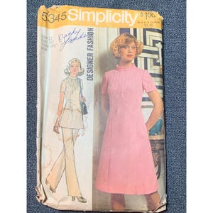May include: Vintage Simplicity sewing pattern number 5345 for a dress and pants. The pattern is for sizes 12-14-16-18. The pattern is for a miss and features a pink dress with a high neck and short sleeves. The dress has a pleated detail at the front. The pants are white and have a wide leg.