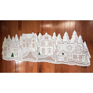 May include: White lace table runner featuring a winter village scene with houses, trees, and street lamps. The intricate design includes detailed windows, doors, and decorative elements, creating a festive holiday aesthetic. The runner is approximately 60 inches long.