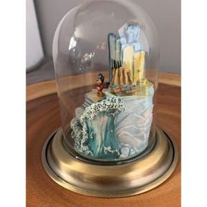 May include: A glass dome with a gold base encloses a miniature scene featuring Mickey Mouse standing on a platform with a wave crashing behind him. The scene is set against a backdrop of a blue and yellow building.