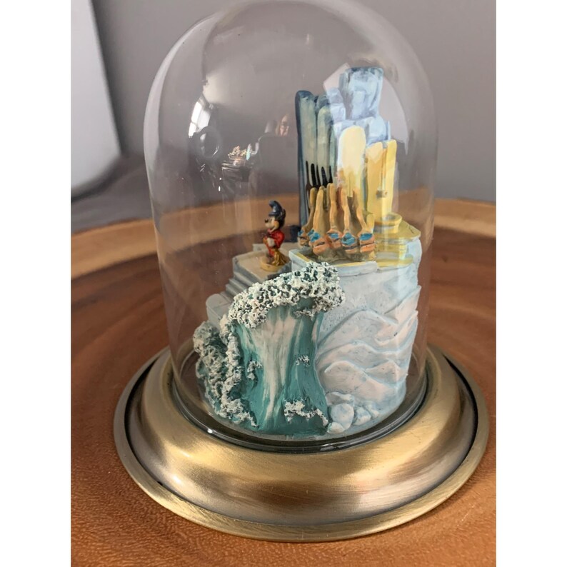 May include: A glass dome with a gold base encloses a miniature scene featuring Mickey Mouse standing on a platform with a wave crashing behind him. The scene is set against a backdrop of a blue and yellow building.
