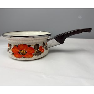 Op de afbeelding: Een crèmekleurige, gespikkelde emaille steelpan met een levendig oranje bloemmotief en bruine bladeren aan de buitenkant. De pan heeft een zilverkleurige metalen rand en een donkerbruine handgreep bevestigd met een gepolijst metalen verbindingsstuk.