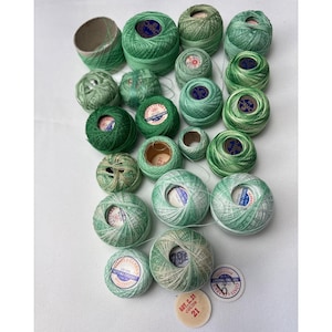 May include: An assortment of green and white thread spools and bobbins. The spools vary in size and shade, from dark forest green to pale mint. Some spools have labels with text, and there is a cardboard tube and a color swatch.