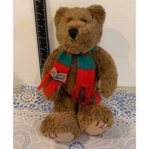May include: A brown plush teddy bear wearing a red and green scarf. The scarf has the word "Bearwear" on it. The bear has a black nose and tan paws. The bear is sitting on a blue and white lace tablecloth.