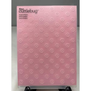 May include: A pink Cricut Cuttlebug embossing folder with a repeating pattern of small flowers. The folder has the text "Cricut cuttlebug" at the top, with "Simple Flowers" and "Fleurs simples" below it. The embossing folder is designed for crafting.