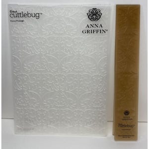 May include: A clear rectangular Cricut Cuttlebug embossing folder and a narrow tan border folder, both featuring the Fancy Plumage pattern. The clear folder displays the text Cricut cuttlebug, Fancy Plumage, and Anna Griffin with a logo.