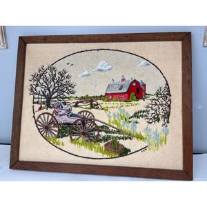 May include: Framed embroidery depicting a rural scene. A red barn with a gray roof is in the background. A white carriage is in the foreground, with a tree on the left and flowers on the right. The embroidery is in a brown wooden frame.
