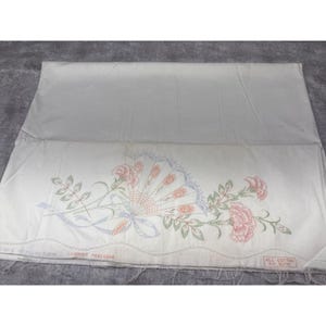 May include: White fabric with a floral design featuring pink carnations, a fan, and blue ribbons. The fabric has the text "COLORTER PROCESSED" and "ALL COTTON" printed on it. The fabric is folded and appears to be a pillowcase.