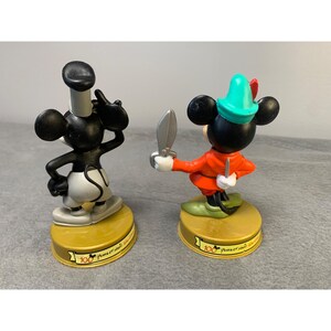 K&ouml;nnte beinhalten: Zwei schwarz-wei&szlig;e Mickey-Maus-Figuren, eine als Seemann und die andere als Robin Hood verkleidet. Beide Figuren stehen auf goldenen Sockeln mit dem aufgedruckten Text "100 Jahre Magie".