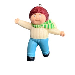May include: A plastic Christmas ornament of a Cabbage Patch Kid wearing a red knit hat, a green and white striped scarf, a white sweater, and blue jeans.