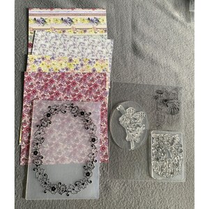 Puede incluir: Un conjunto de materiales para manualidades que incluye una pila de hojas de papel con diseños florales y de enredaderas, una carpeta de relieve de plástico transparente con una corona de flores ovalada y dos sellos de silicona.