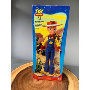 Może przedstawiać: Tekturowe pudełko na lalkę Jessie z filmu Toy Story 2 produkcji Disney Pixar. Pudełko przedstawia Jessie w ż&oacute;łtej koszulce, niebieskich ogrodniczkach i brązowym kowbojskim kapeluszu. Tekst na pudełku brzmi: "Najlepsze przyjaci&oacute;łki na zawsze Jessie i ty. Ona wie, że ją kochasz i ona też cię kocha!"