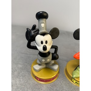 K&ouml;nnte beinhalten: Eine schwarz-wei&szlig;e Mickey Mouse Figur, die einen grauen Matrosenhut tr&auml;gt. Die Figur steht auf einem gelben Sockel mit dem aufgedruckten Text "Steamboat Willie".