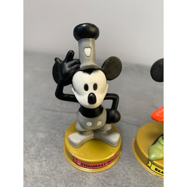 K&ouml;nnte beinhalten: Eine schwarz-wei&szlig;e Mickey Mouse Figur, die einen grauen Matrosenhut tr&auml;gt. Die Figur steht auf einem gelben Sockel mit dem aufgedruckten Text "Steamboat Willie".
