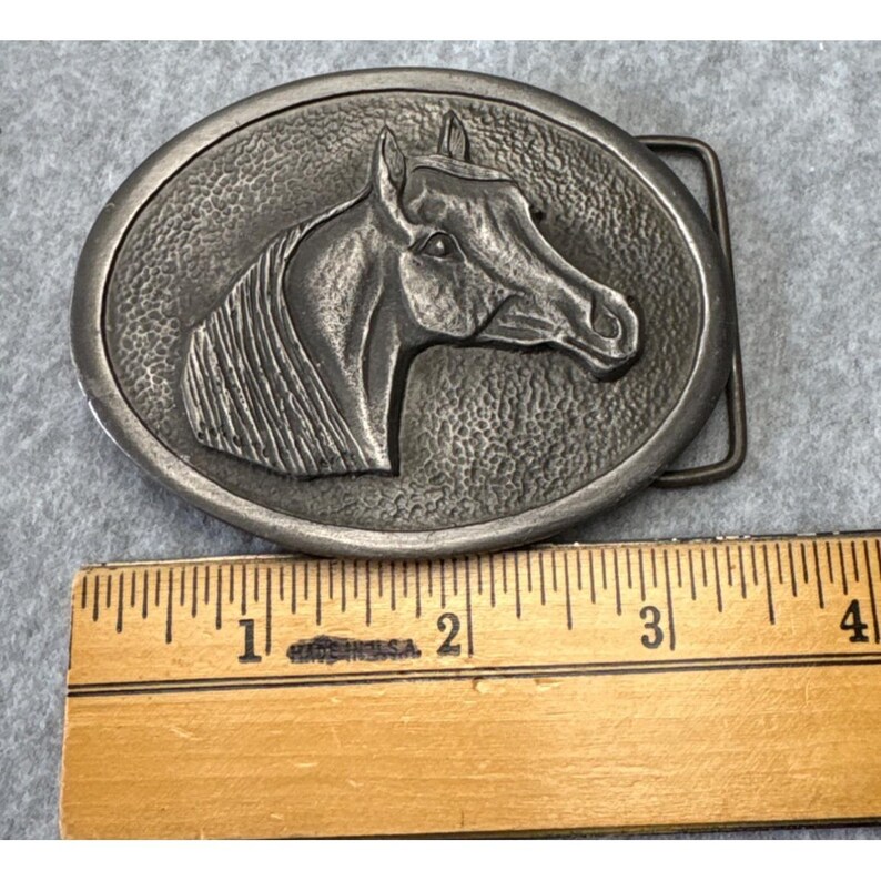 May include: Oval-shaped, silver-toned metal belt buckle featuring a detailed relief of a horse's head. The horse's profile is centered within a textured border. The buckle includes a standard belt loop and pin. The ruler shows the buckle is approximately 4 inches long.