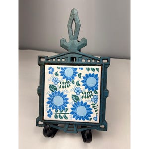 May include: A teal metal trivet with a square tile insert. The tile features a floral pattern with blue flowers and green leaves on a white background. The trivet has a handle and small wheels.