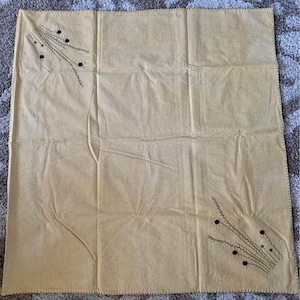 May include: A square, light tan fabric with decorative black embroidered floral designs in two corners. The fabric has a stitched border and visible creases. The floral designs consist of thin stems, zigzag lines, and small black circles.