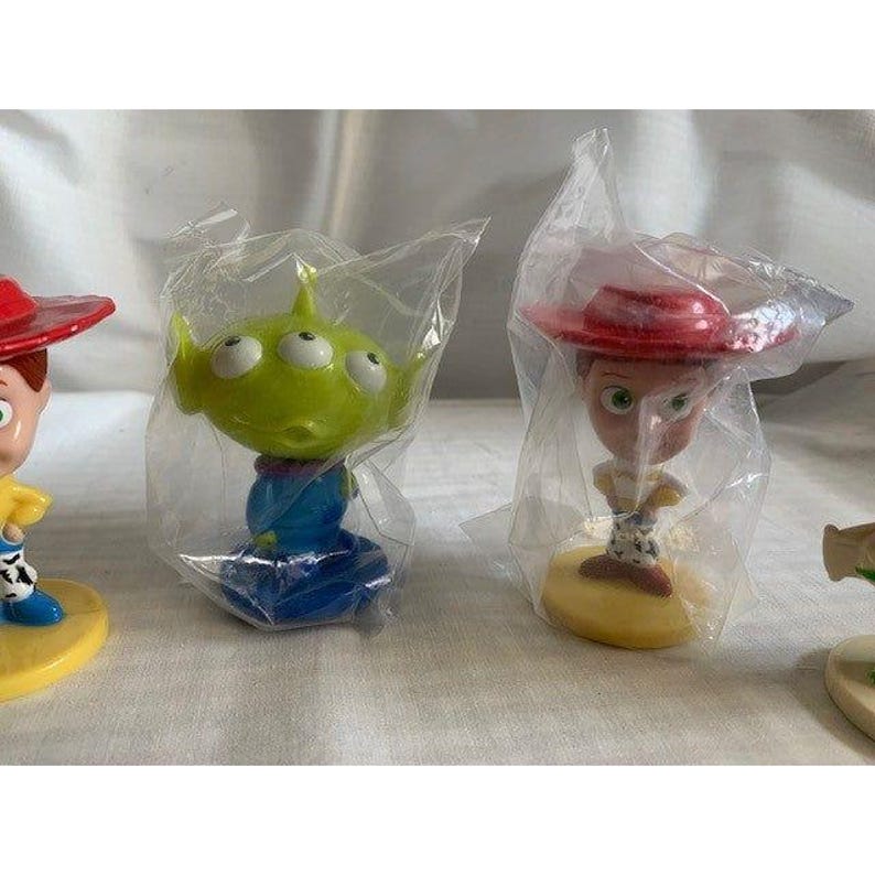 May include: Several small character figurines from the Toy Story franchise, including Woody, an alien, and Jessie, are visible. Each figure is in a clear plastic bag. The figures have bright colors and are on a white surface.