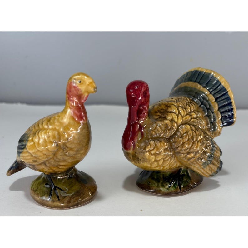 May include: A pair of ceramic turkey figurines. One turkey is standing upright, and the other is in a more relaxed pose. The turkeys are painted in shades of yellow, brown, red, and blue. They are likely decorative items.