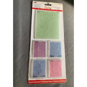 May include: A Sizzix Textured Impressions embossing folder set featuring one large light green folder with a swirl pattern and four smaller folders in pink and blue with designs including the words LOVE, FOREVER, bells, and a flower.