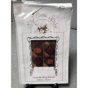 May include: A package featuring a craft pattern for a November Bitty Banner. The banner design includes a decorative metal frame holding a patchwork of autumn-themed squares with pumpkins and leaves. The package has the text "Lazy Anna Stitches".