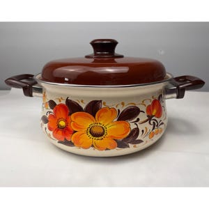 May include: A round, cream-colored enamel cooking pot with a matching brown lid. The pot features a decorative floral pattern with large orange and yellow flowers, dark brown leaves, and gold scroll accents. It has two brown side handles.