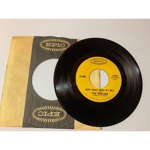 May include: A black vinyl record with a yellow label featuring the text "NOT THAT WAY AT ALL" by The Hollies. The record is next to its tan and black paper sleeve, which has the Epic Records logo.
