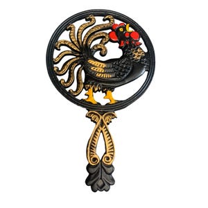May include: A cast iron trivet featuring a rooster design. The trivet is black with gold and yellow accents. The rooster is inside a circular frame with a decorative handle. The trivet measures approximately 8 inches in diameter.