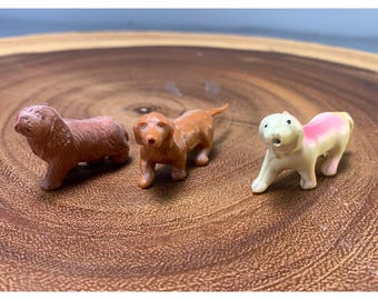 May include: Three miniature dog figurines in shades of brown and pink are arranged on a wooden surface. The dogs vary in breed and pose, with one appearing to be a dachshund. The figurines are small and detailed.