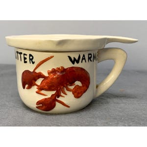 May include: White ceramic butter warmer with a lobster design and the words "Butter Warmer" on the side. The warmer has a lid with a small handle.