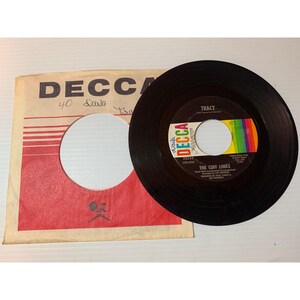 May include: A black 45 RPM vinyl record by The Cuff Links titled Tracy on the Decca label, featuring a central black label with white and colored text. It is accompanied by a square paper sleeve with the Decca logo and red graphic accents.