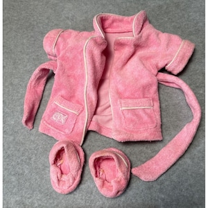 May include: A pink terry cloth robe set with white trim, including a short-sleeved robe with pockets and a matching belt, and two pairs of slippers. The robe has a logo on the pocket.