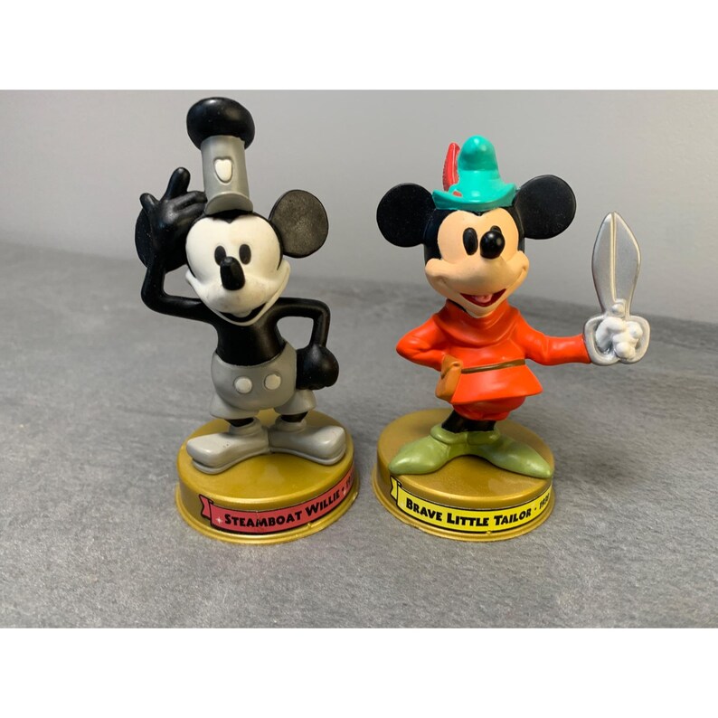 K&ouml;nnte beinhalten: Zwei Vintage-Plastikfiguren von Mickey Mouse. Die Figur links ist als Steamboat Willie mit einem schwarz-wei&szlig;en Matrosenanzug und einem schwarzen Hut gekleidet. Die Figur rechts ist als der Brave Little Tailor mit einem rot-gr&uuml;nen Outfit und einem gr&uuml;nen Hut gekleidet. Beide Figuren stehen auf einem runden Sockel mit dem Namen des Charakters darauf.