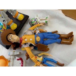 May include: Several Toy Story character action figures, including Woody, Buzz Lightyear, and others. Woody is wearing a yellow plaid shirt, blue pants, and brown boots. Buzz Lightyear is in a white and green spacesuit. Other figures are also visible.
