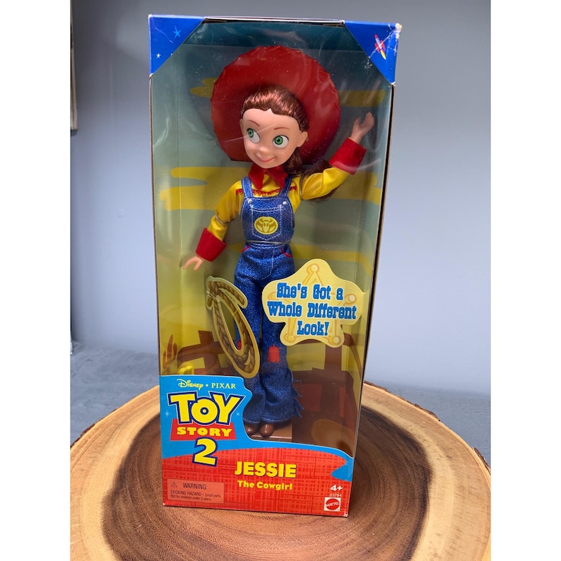 Może przedstawiać: Lalka Jessie Kowbojki z Toy Story 2 w pudełku. Lalka ma na sobie czerwony kapelusz, ż&oacute;łtą koszulę, niebieskie ogrodniczki i brązowe buty. Pudełko ma na przodzie zdjęcie Jessie i napis "She's Got a Whole Different Look!" u dołu. Pudełko ma r&oacute;wnież logo Toy Story 2 i napis "Jessie The Cowgirl" na przodzie.