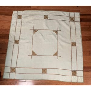 May include: A light green tablecloth with a square design in the center and decorative squares in each corner. The tablecloth has a brown embroidered border and accents. The fabric is smooth and appears to be made of a soft material.