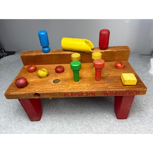May include: A vintage Playskool workbench toy made of wood, featuring a brown tabletop and red legs. The workbench includes colorful wooden pegs, screws, and a yellow hammer. The text "Playskool Workbench" is printed on the front.