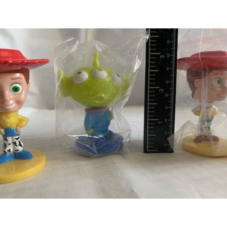 May include: Three Toy Story character figurines, including Woody and an alien, are displayed. The figures are in clear plastic packaging. A black ruler with white markings is positioned between the figures.