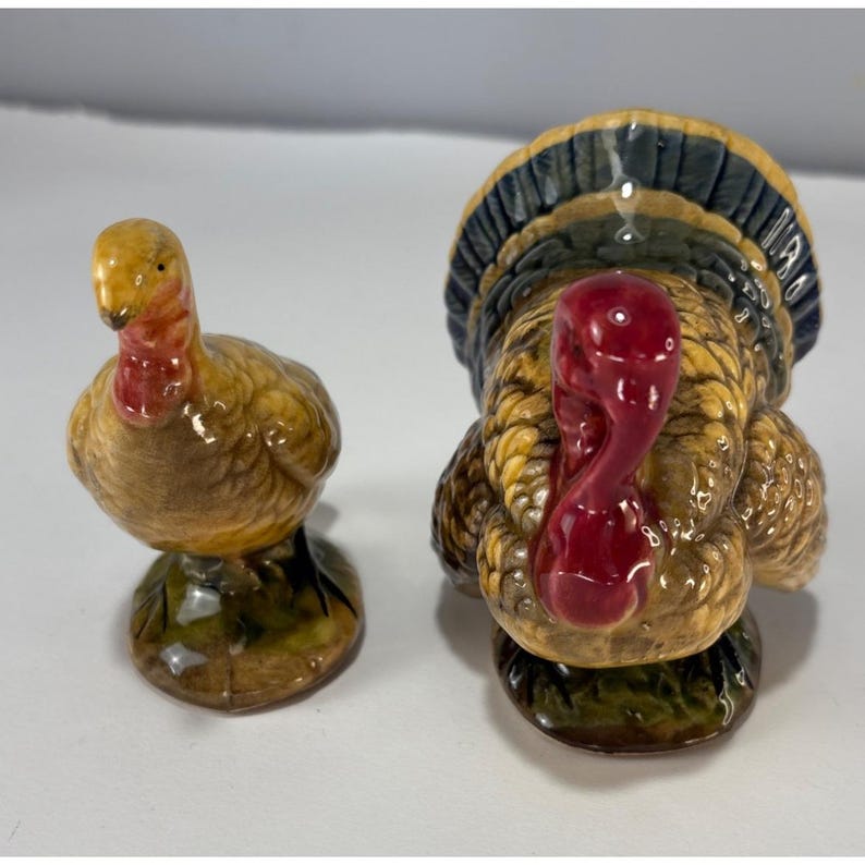 May include: A pair of ceramic turkey figurines. One is a hen, the other a tom. The hen is small and round, with a yellow head and red wattle. The tom has a large, fanned tail and a red wattle. Both are on green bases.