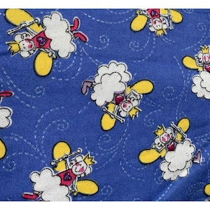 May include: Blue fabric with a repeating pattern of cartoon sheep wearing crowns and holding wands. The sheep have white fluffy bodies, yellow wings, and pink accents. The background has a subtle swirl design.