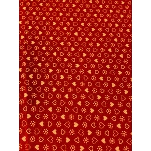 May include: Red fabric with a repeating pattern of small, light orange hearts and floral-like shapes. The hearts and floral shapes are evenly spaced across the fabric, creating a consistent design. The fabric has a smooth texture.