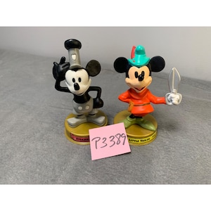 K&ouml;nnte beinhalten: Zwei Vintage-Mickey-Mouse-Figuren, eine als Dampfschiffkapit&auml;n und die andere als kleiner Schneider verkleidet. Beide Figuren sind schwarz-wei&szlig; mit roten Akzenten und stehen auf einem runden Sockel.