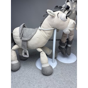 K&ouml;nnte beinhalten: Ein Pl&uuml;schpferd in Hellbeige mit grauem Sattel, M&auml;hne und Hufen. Das Pferd hat ein skurriles Design mit einem l&auml;chelnden Gesicht und einem wei&szlig;en Metallst&auml;nder. Das Spielzeug ist als Schaukelpferd konzipiert.
