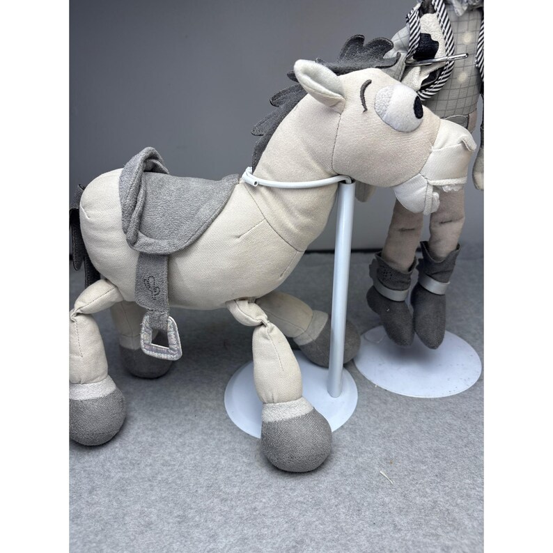 K&ouml;nnte beinhalten: Ein Pl&uuml;schpferd in Hellbeige mit grauem Sattel, M&auml;hne und Hufen. Das Pferd hat ein skurriles Design mit einem l&auml;chelnden Gesicht und einem wei&szlig;en Metallst&auml;nder. Das Spielzeug ist als Schaukelpferd konzipiert.