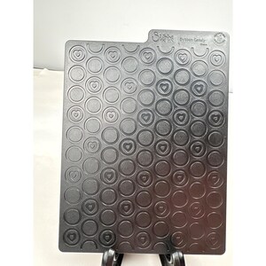 May include: A dark gray Sizzix die-cutting plate with a pattern of circles and hearts. The plate has a rectangular shape with a small tab on the top right corner. The words "Sizzix" and "Burton Candy" are printed on the plate.