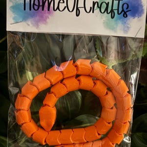 May include: A bright orange articulated snake toy made of connected segments, packaged in a clear plastic bag with a white header card featuring the text HomeOfCrafts in black script with colourful watercolour-style accents on the edges.