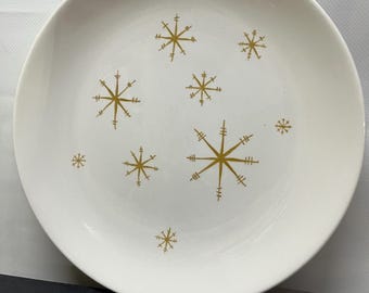 May include: A white ceramic plate with a gold snowflake design. The plate has a simple, elegant design with various sizes of snowflakes scattered across the surface. The snowflakes are a warm gold color, contrasting against the white background.